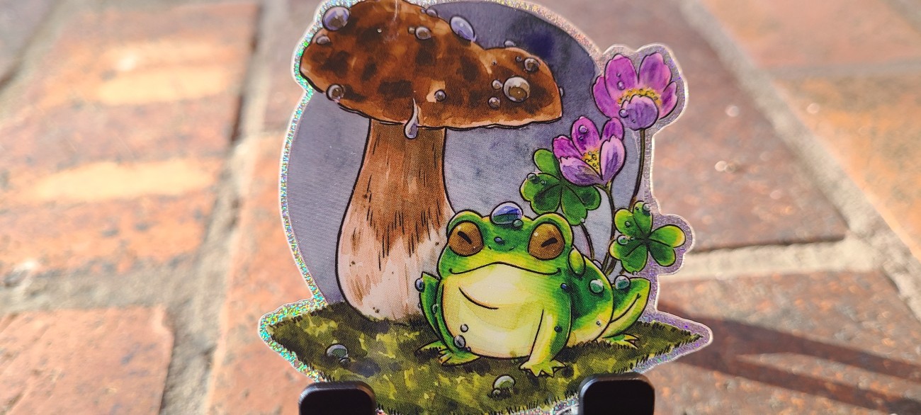 A glittery vinyl sticker of a little green frog happily sitting beneath a mushroom.  There are raindrops on the frog, mushroom, and on the sorrel flowers behind.